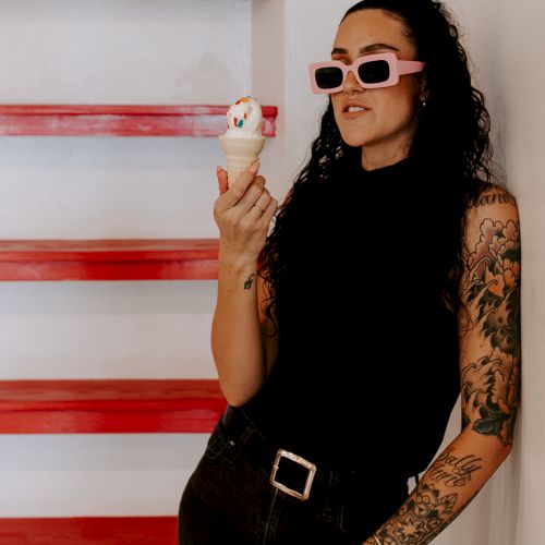 A person with tattoos, wearing sunglasses, is holding an ice cream cone and standing by red shelves.
