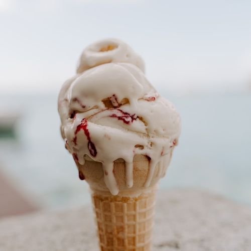 An ice cream cone with swirled, melting ice cream and red syrup, set against a blurred outdoor background with water in the distance.