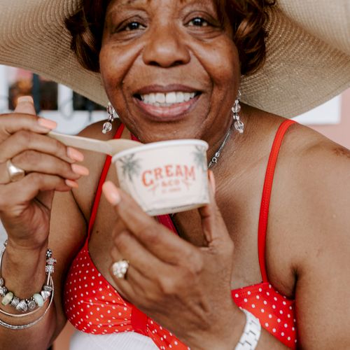 A woman in a sunhat and polka dot dress is smiling, holding a small cup of cream.