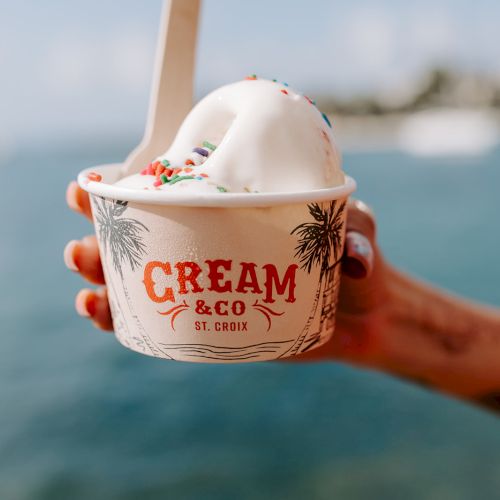 A hand holds a cup of ice cream with a spoon, branded "Cream & Co St. Croix," against a blurry seaside background.