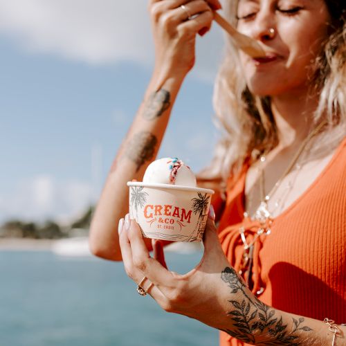 A person enjoys ice cream by the water, holding a cup labeled "CREAM." They're wearing an orange top with visible tattoos.