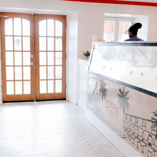 The image shows an ice cream shop with a counter and display, wooden doors, a stool, and a person standing behind the counter.