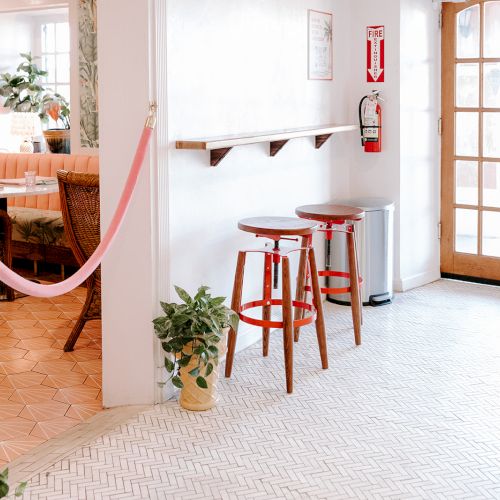 The image shows a bright space with stools, plants, a French door, and a pink rope divider separating a dining area with chairs and a table.