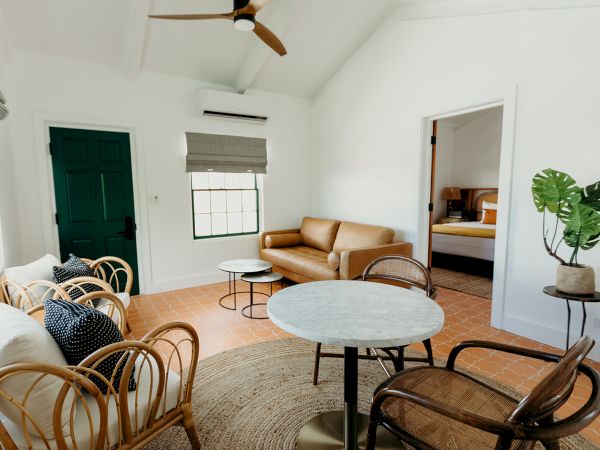 A cozy, modern room with wicker chairs, a sofa, round tables, and a plant; leads to a bedroom with white walls and terracotta floors.