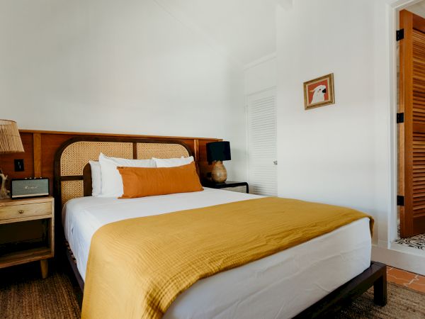 A cozy bedroom with a bed, white and mustard bedding, bedside tables, lamps, and a partially open door to another room.