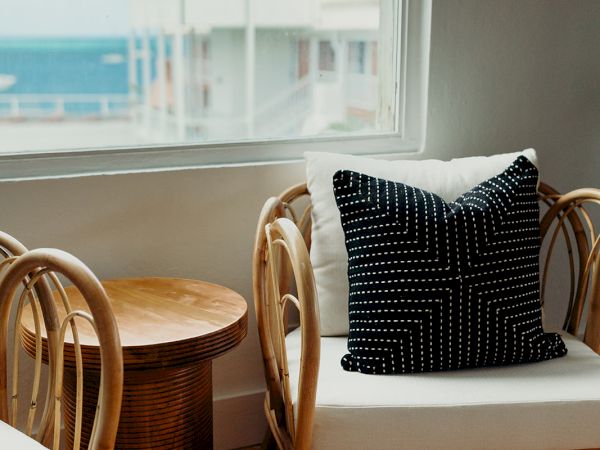 The image shows a cozy sitting area with wicker chairs, a patterned cushion, a small wooden table, and a seaside view through a window.
