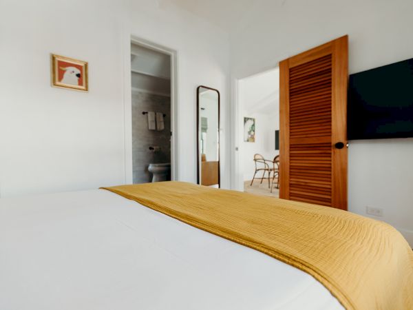 A bedroom with a bed, yellow blanket, a TV, and a wooden door; artwork and a mirror are on the walls, with a view into the bathroom.