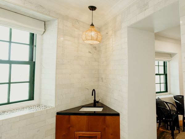 A small corner sink with a wooden cabinet, pendant light, and patterned tile floor in a room with brick walls and a window.