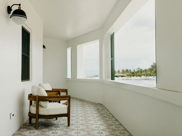 A balcony with two wooden chairs, white walls, patterned tile flooring, overlooking a scenic view of the ocean and trees in the distance.