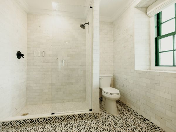 The image shows a bathroom with a glass-enclosed shower, patterned tile floor, white brick walls, a toilet, and a window with green trim.