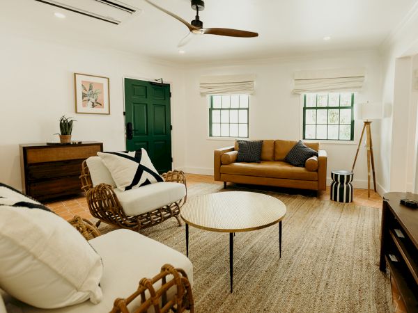 A cozy living room with modern decor, featuring a brown sofa, wicker chairs, a round table, a green door, and natural light.