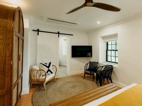 A cozy room with a bed, wicker chairs, a wooden wardrobe, TV, ceiling fan, and a sliding door leading to a bathroom.