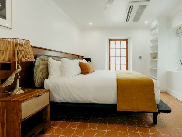 A cozy bedroom with a large bed, white and mustard bedding, a wooden nightstand with a lamp, open shelves, and a small plant by the window.