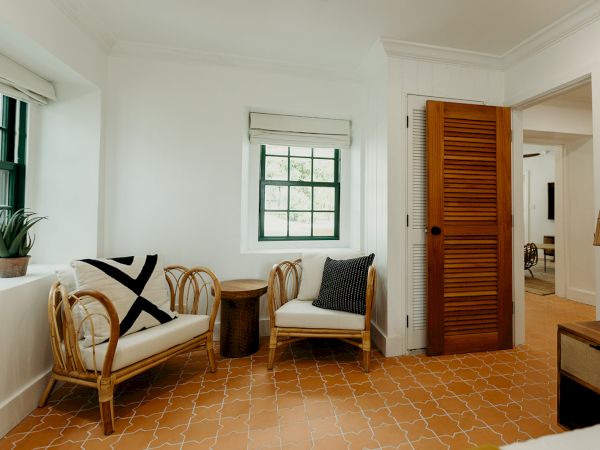 A cozy room with two wicker chairs, cushions, a small table, plant, and terracotta tiled floor. Doorway leads to another room.