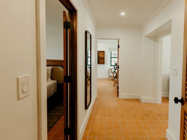 A hallway with tile flooring, leading to a bedroom and living area with wooden furniture and white walls, captured in warm lighting.