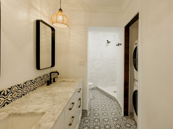 A bathroom with a marble countertop, patterned tiles, a shower, and a stacked washer and dryer combo unit in the corner.