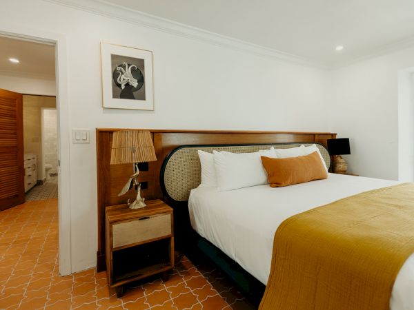 A cozy bedroom with a large bed, orange blanket, wooden headboard, beside lamps, and open door to a bathroom, in a warmly lit space.