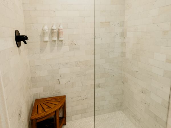 A modern shower with white tiles, a glass door, black fixtures, a wooden corner stool, and a few bottles on a wall rack.