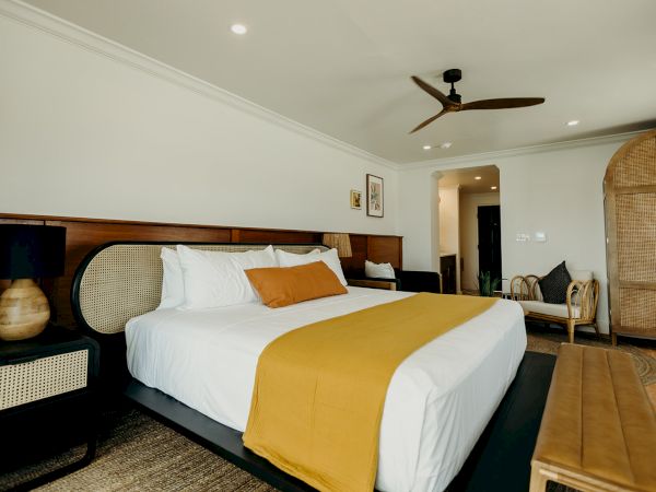 A modern bedroom with a large bed, yellow accents, wooden furniture, and ceiling fan. Cozy and inviting atmosphere.