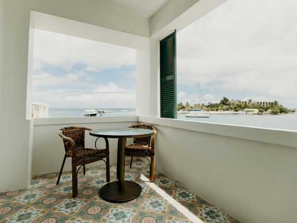 A small balcony with two wicker chairs and a round table overlooking the sea. There’s a tiled floor and a view of distant boats and an island.