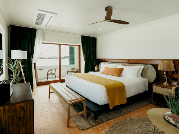 A cozy bedroom with a large bed, ceiling fan, plants, and a view through the sliding glass door, creating a warm and inviting atmosphere.