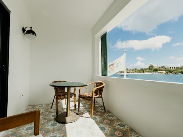 A small balcony with two chairs and a table, overlooking a body of water and distant buildings, with a pleasant, sunny sky.