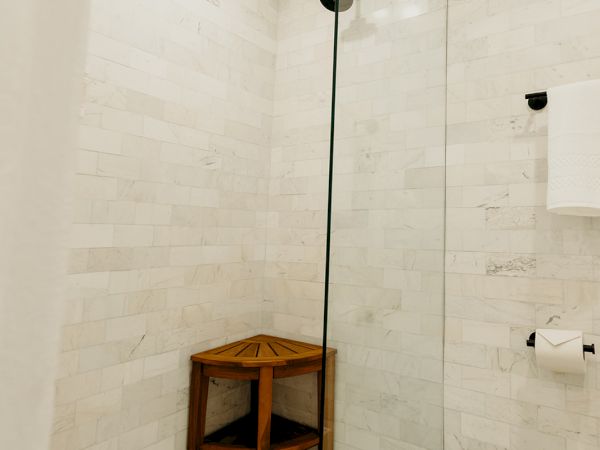 A modern shower with white tiles, a glass door, a large rainfall showerhead, and a small wooden bench in the corner.