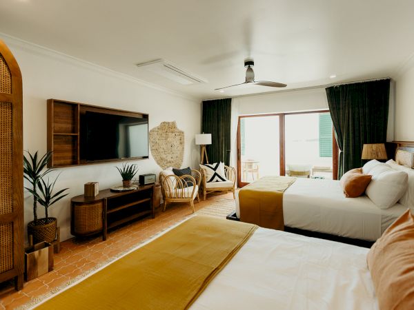 A cozy bedroom with two beds, TV, plants, and wicker accents, enhanced by natural light from a large window, creating a warm ambiance.