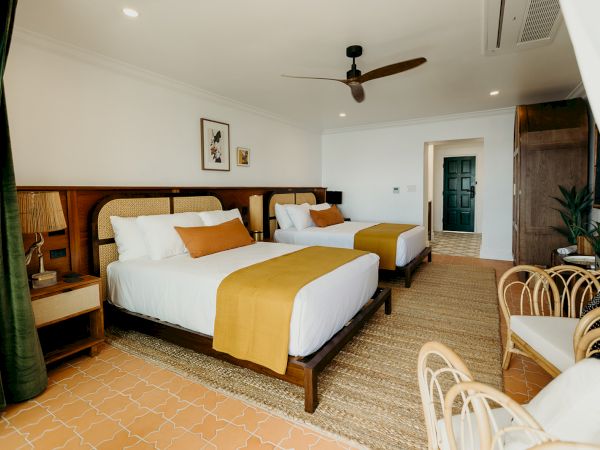 The image shows a stylish hotel room with two double beds, modern decor, a ceiling fan, and rattan furniture, creating a cozy atmosphere.