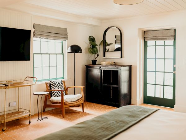A cozy room with a large window, armchair, TV, and a cart. Features a dark cabinet with a mirror and plants, and a door with glass panels.