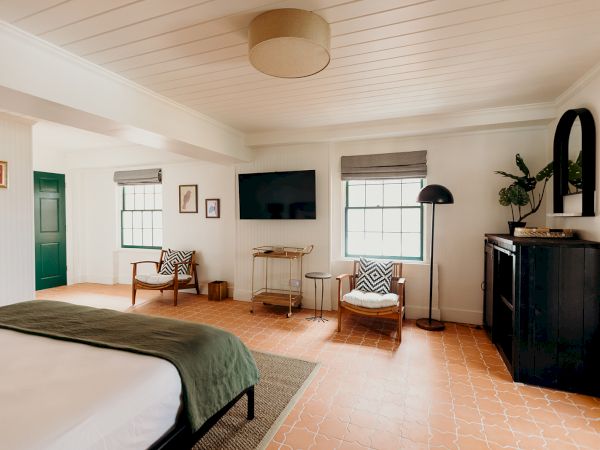 A cozy bedroom with a bed, TV, armchairs, and decor on light-colored walls and floor.