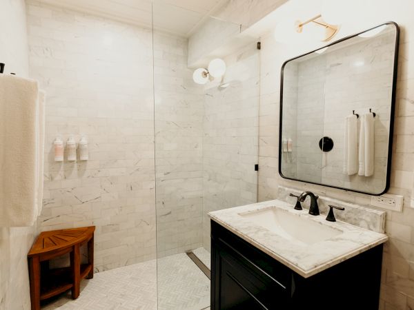 A modern bathroom with a glass shower, marble sink, large mirror, and towels. There’s a wooden bench and toiletries in the shower area.