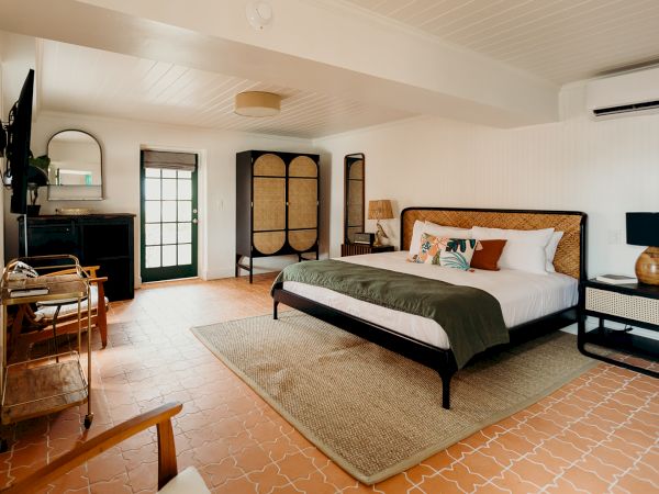 A cozy bedroom with a large bed, TV, chairs, wardrobe, and a rug on tiled flooring. The room features natural tones and a door to outside.