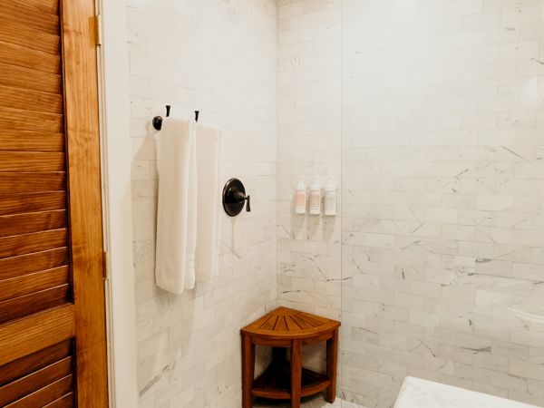 A bathroom featuring a wooden door, white tiled walls, a towel on a rack, a wooden stool, and decorative patterned floor tiles.