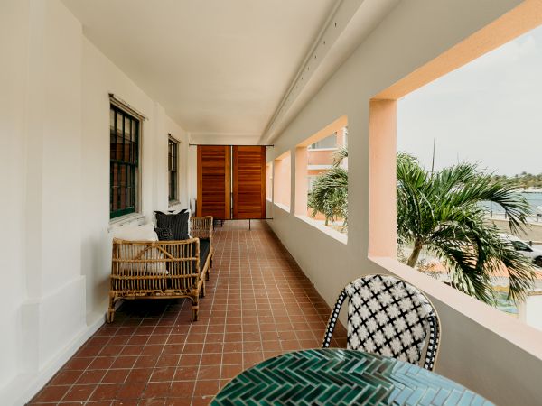 A veranda with wicker furniture, a table, and chairs overlooks a palm tree and waterfront view, with white walls and tile flooring.