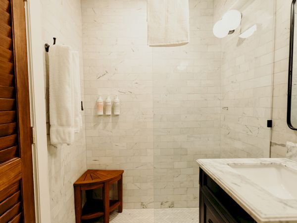 A small bathroom featuring a walk-in shower, wooden bench, marble counter, wall-mounted lights, and decorative floor tiles.