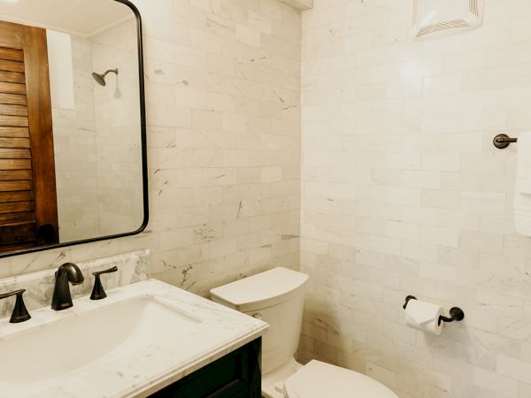 A bathroom with a toilet, a black vanity with a sink, a mirror, and patterned floor tiles. The walls are light-colored tiles.