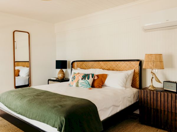 A cozy bedroom with a double bed, green blanket, decorative pillows, and wooden side tables. A mirror and air conditioner are on the wall.