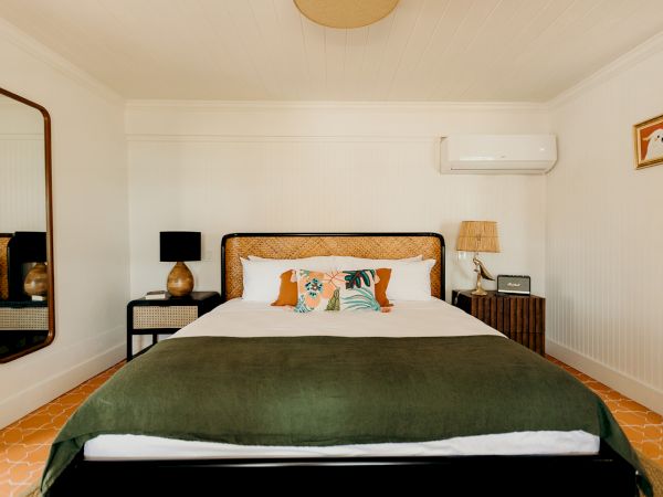 A bedroom with a neatly made bed, green blanket, decorative pillows, bedside tables, lamps, wall art, mirror, and an air conditioner on white walls.