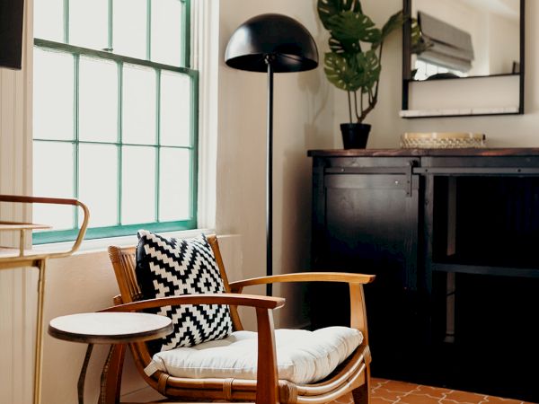 A cozy room with a wooden chair, geometric pillow, small table, black cabinet, tall lamp, plant, and window with Roman shade, under soft lighting.