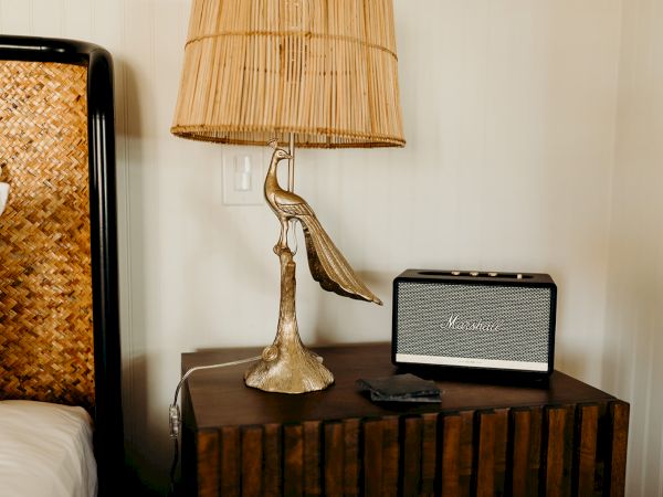 A decorative lamp with a bird base and a speaker are on a wooden bedside table next to a woven headboard.