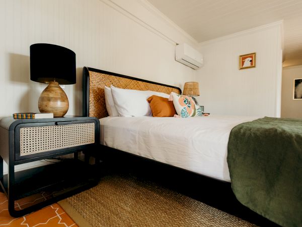 A cozy bedroom featuring a bed with pillows, a bedside table with a lamp and books, and a green blanket on a woven rug.