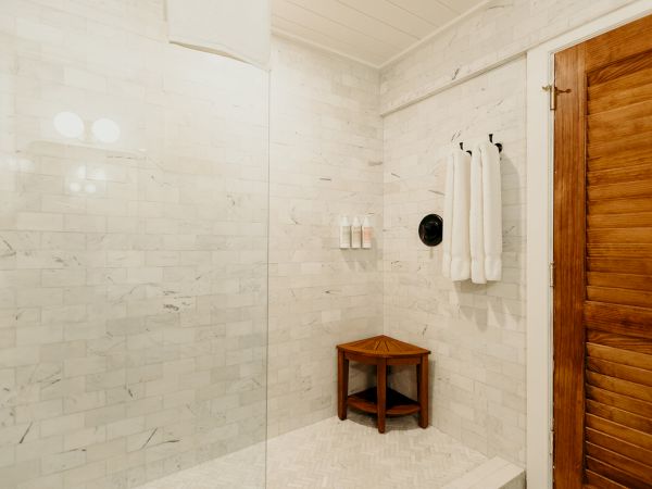 The image shows a shower with white tiled walls, a wooden bench, towels, and a glass divider, adjacent to a wooden door.