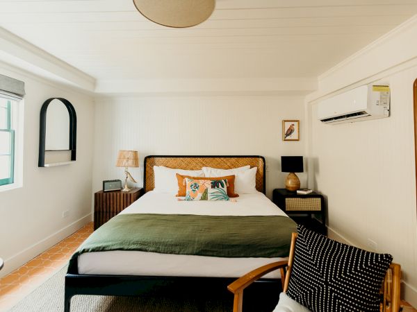 A cozy bedroom with a bed, stylish lamps, a chair, and a wall mirror. The room has warm lighting and a modern, minimalist design.