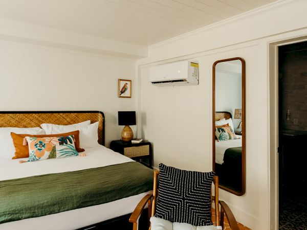 A cozy bedroom with a bed, decorative pillows, a wooden chair, a mirror, and an air conditioner on the wall.