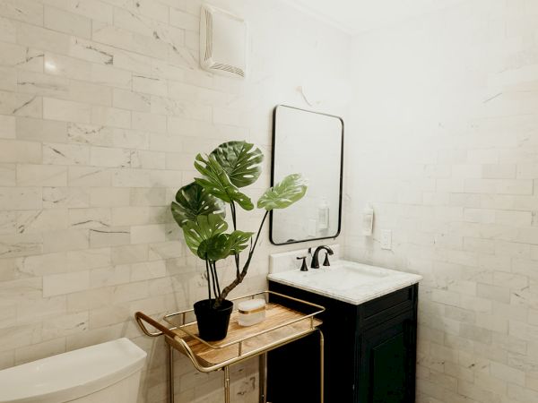 A small bathroom features a toilet, a sink with a mirror, a decorative plant on a wooden cart, and black and white patterned floor tiles.