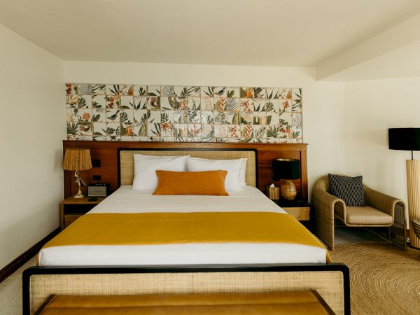 The image shows a bedroom with a large bed, colorful headboard, lamps, a chair, and a rug, in a cozy, well-decorated space.