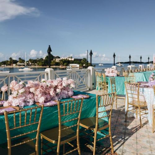 Elegant outdoor dining setup with teal tablecloths, gold chairs, and pink floral centerpieces by the waterfront under a clear blue sky.