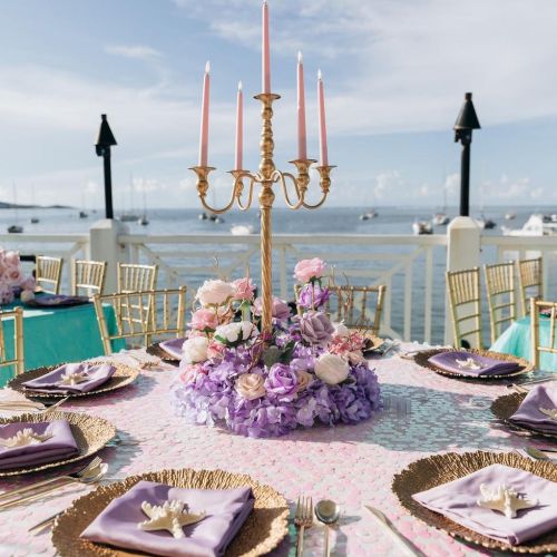 A beautifully set outdoor table by the sea, featuring a purple floral centerpiece with a gold candelabrum, surrounded by elegant tableware.