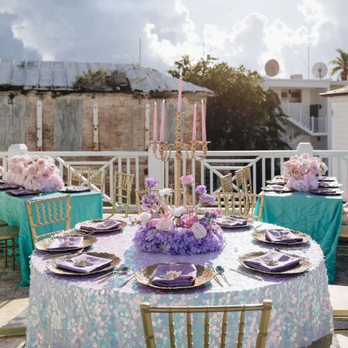 An elegant outdoor table setting with purple and pink floral centerpieces, gold chairs, and decorative plates, under a cloudy sky.